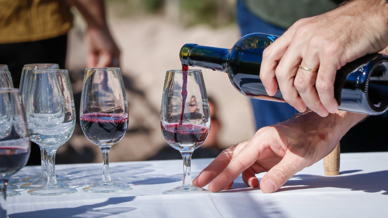 A closeup view of a man’s hand pouring a glass of vegan wine with empty and full glasses next to it outdoors with people in the background